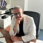 Foto di Mauro Pianezzi, medico ortopedico direttore di OTODI Sardegna, seduto alla sua scrivania con il camice bianco e occhiali blu.